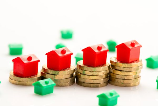 Building Safety Levy 2026: Four red toy houses stacked on rising piles of gold coins, with small green houses scattered around them against a plain white background.