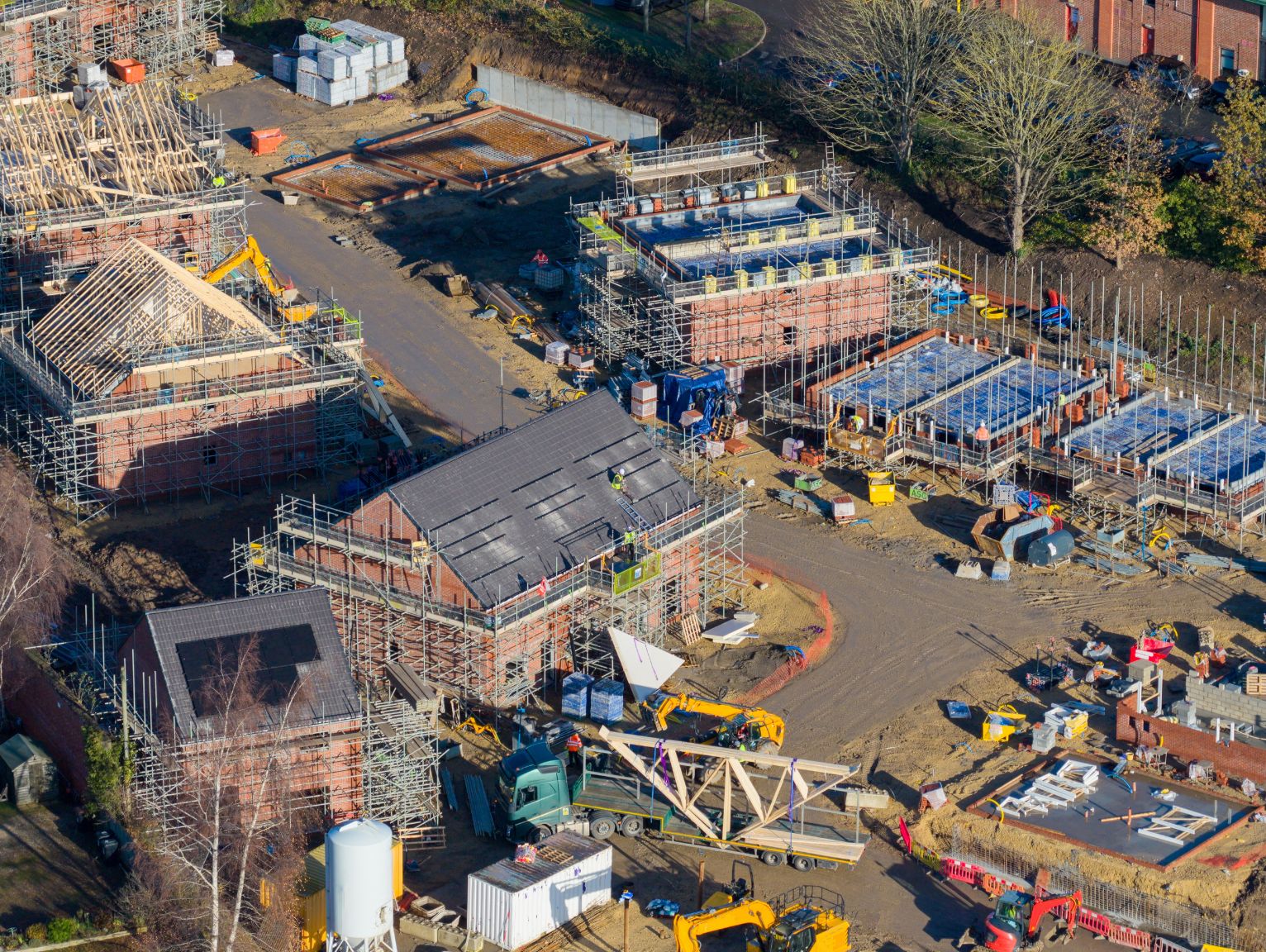 Building safety levy exemptions: An aerial view of a large new-build housing development under construction, showing timber roof trusses, brick shells wrapped in scaffolding, and building materials, vehicles and workers scattered across the muddy site.