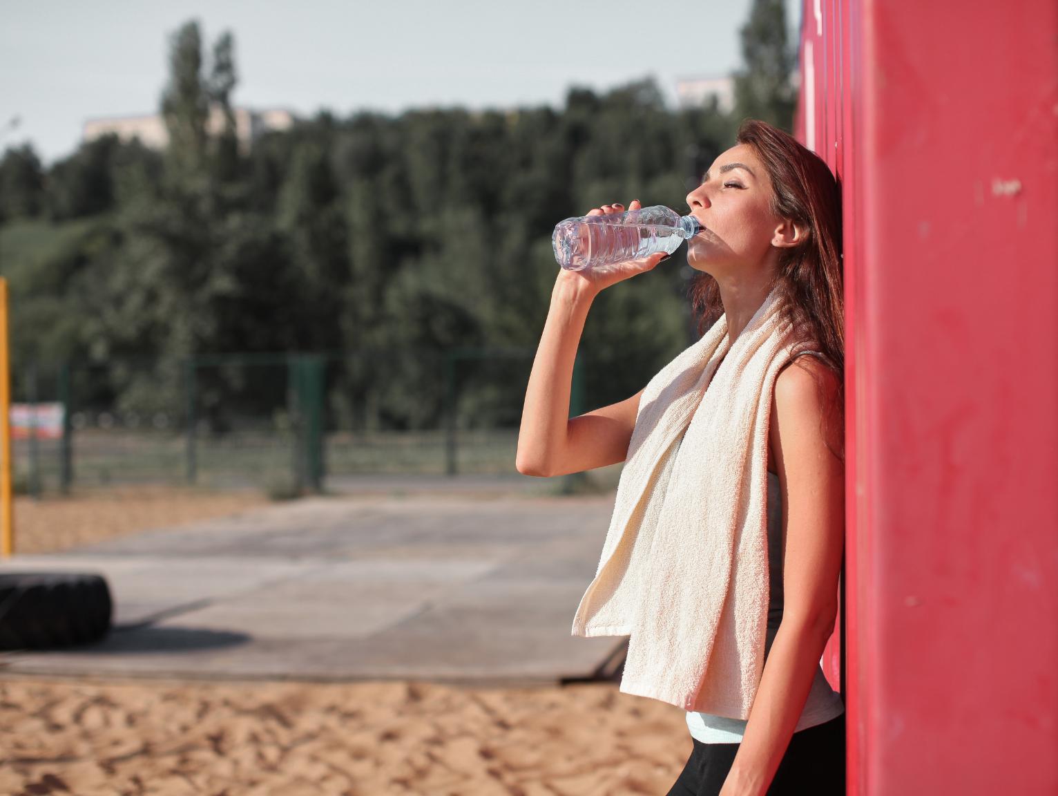 Container gyms: A woman in workout clothes with a towel around her neck drinking from a water bottle while leaning against a red shipping container outdoors.