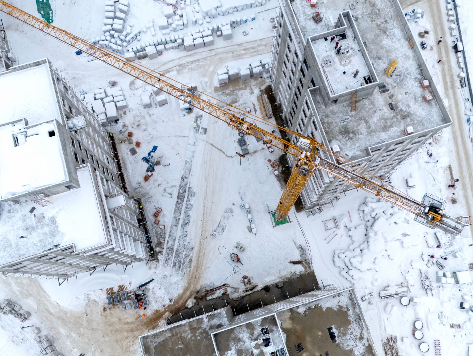 HSE welfare requirements construction site: Aerial view of a snow-covered construction site with a yellow tower crane between unfinished concrete apartment blocks and stacked materials.