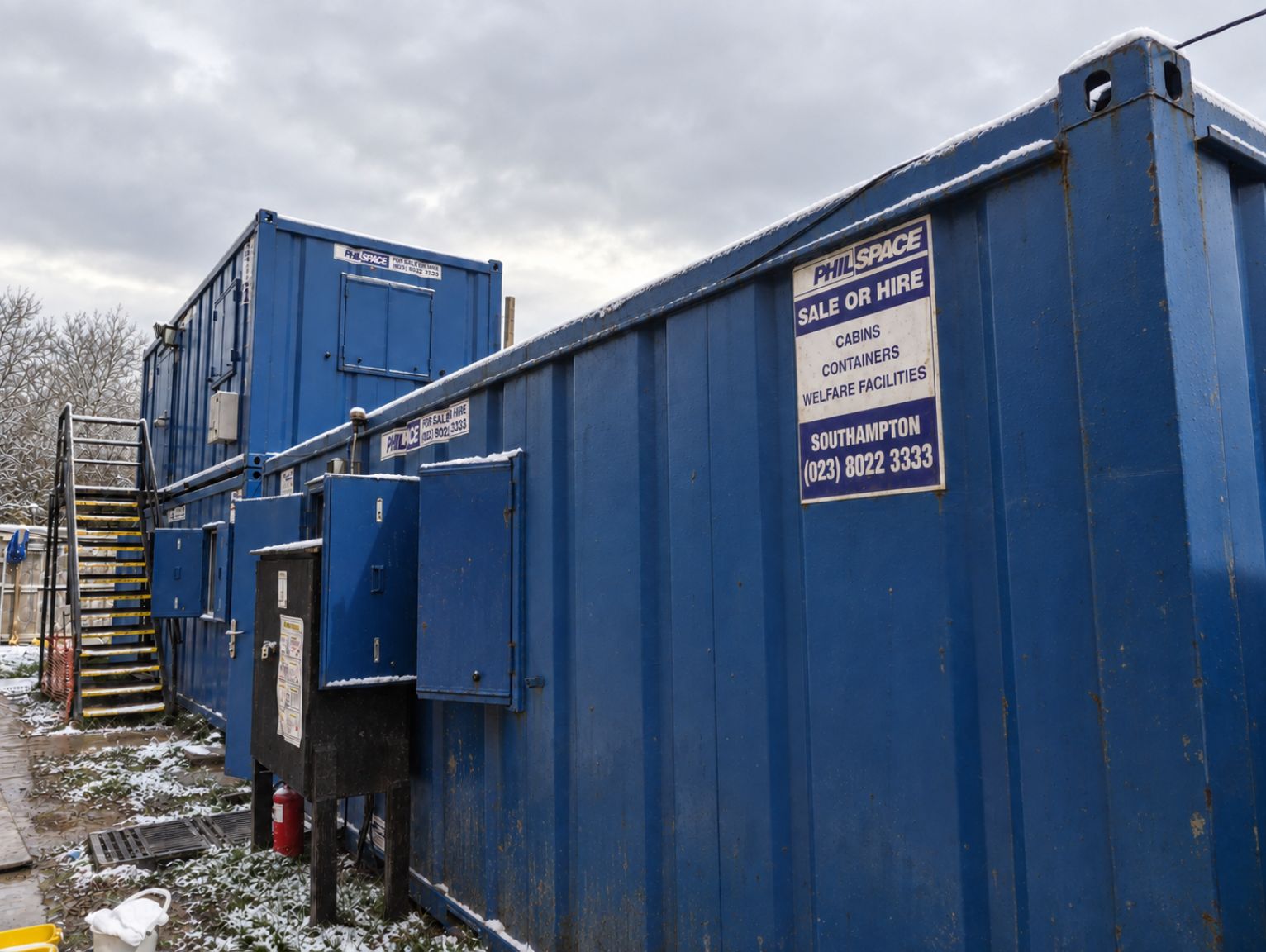 How Many Welfare Units Do I Need On-Site: Row of blue PhilSpace site cabins with “Sale or Hire” signs, external stairs and a light dusting of snow under a grey sky.