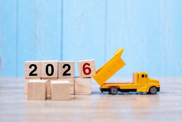How Will the Autumn Budget Affect Construction Costs: Wooden blocks spelling out 2026 on a wooden surface, with a yellow toy tipper truck tilting its raised bed against the red number 6, set against a light blue wooden background.