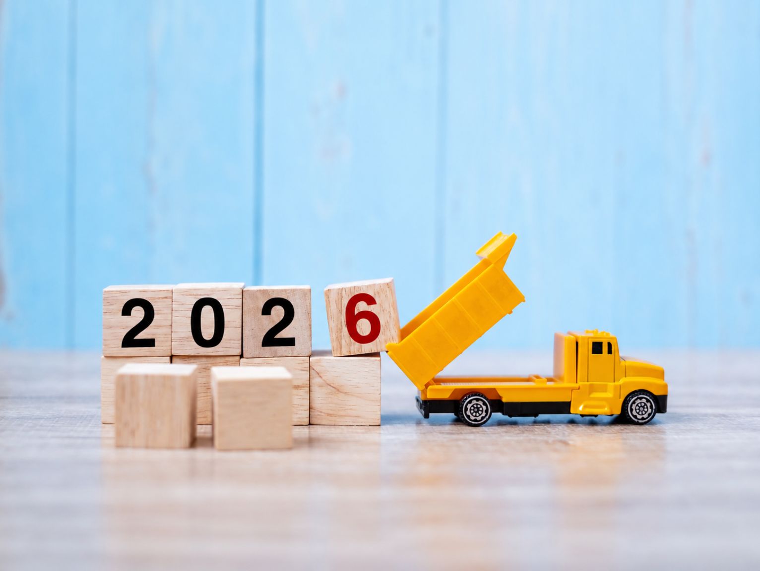 How Will the Autumn Budget Affect Construction Costs: Wooden blocks spelling out 2026 on a wooden surface, with a yellow toy tipper truck tilting its raised bed against the red number 6, set against a light blue wooden background.