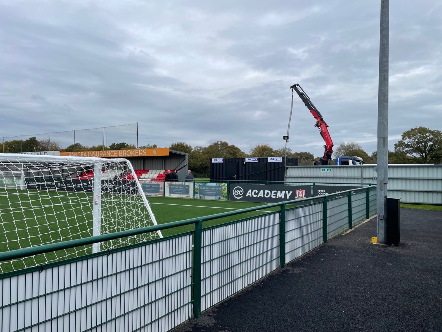 A wide view of Philspace cabins being lifted into position at Sholing FC, with the pitch and stands visible in the background.