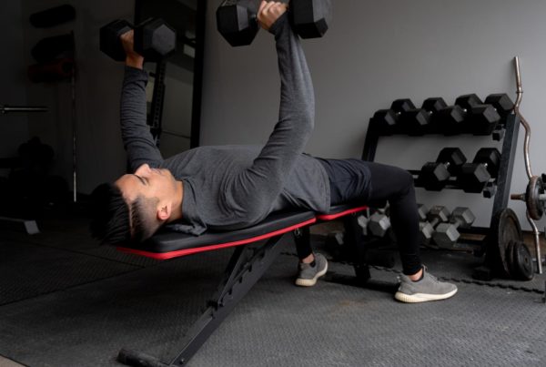Shipping Container Gyms: A man lying on a weight bench performing a dumbbell chest press in a small gym, with a rack of dumbbells visible in the background.