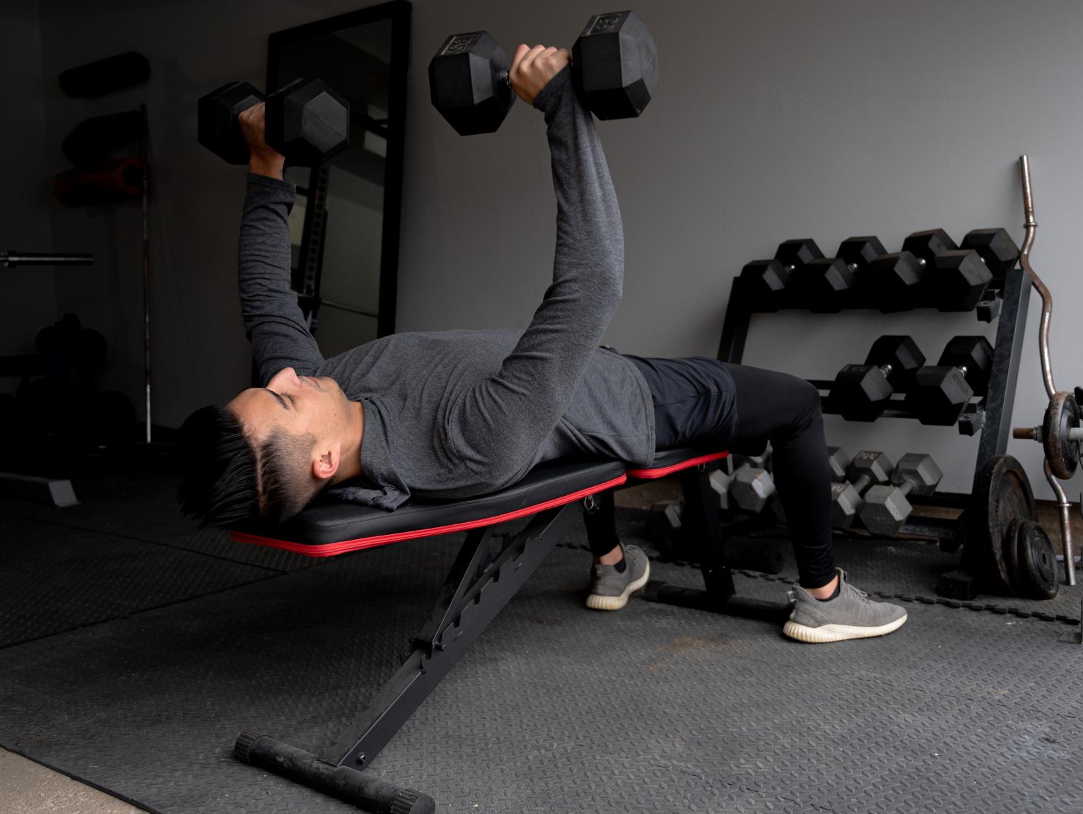 Shipping Container Gyms: A man lying on a weight bench performing a dumbbell chest press in a small gym, with a rack of dumbbells visible in the background.