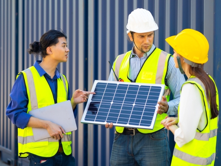 Three people in hi vis jackets and hardhats hold a solar panel by a sustainable blue shipping container