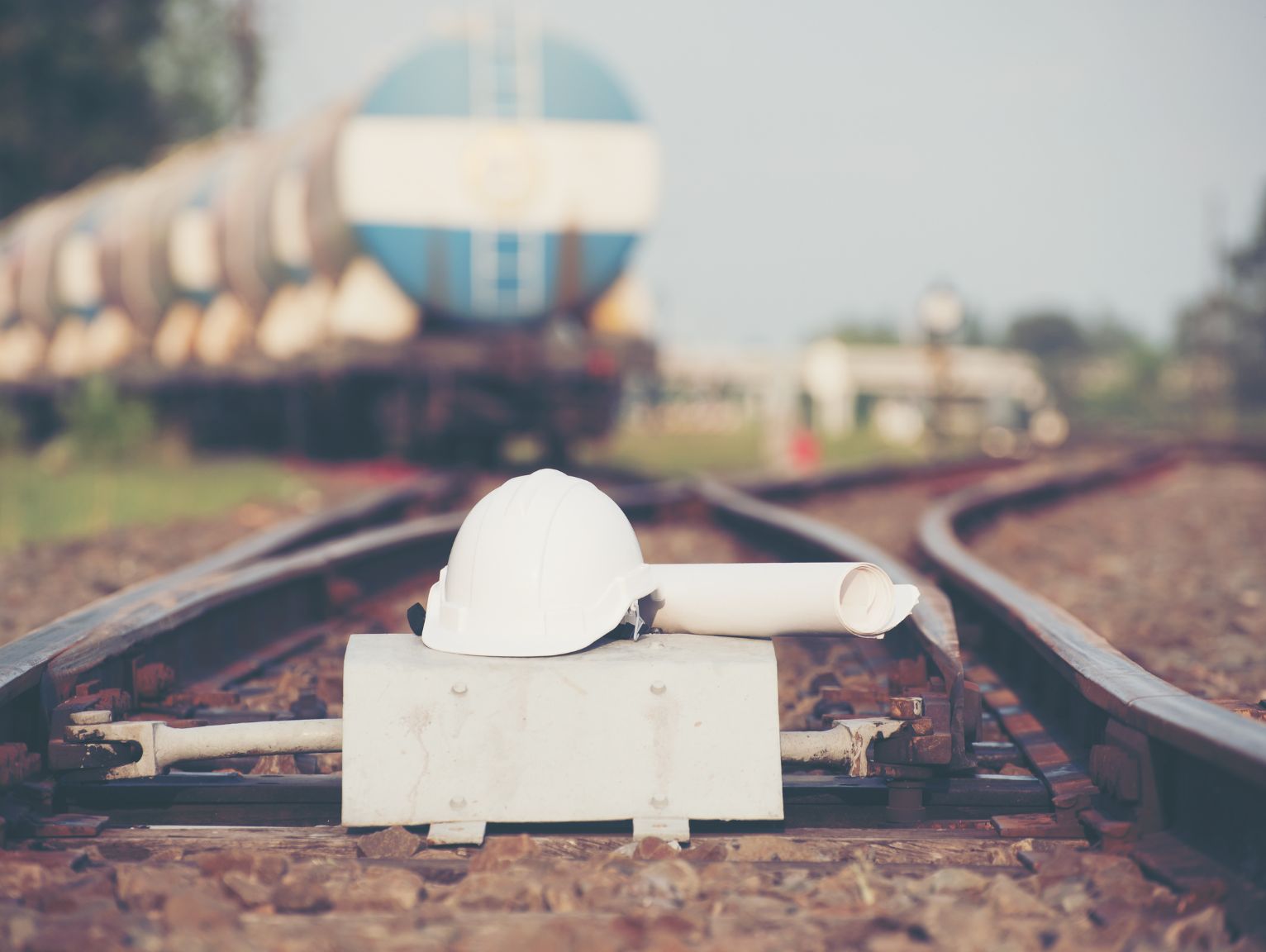 Welfare compliance for mobile infrastructure crews: A white hard hat and rolled-up technical drawings resting on railway track points, with a blurred blue freight train visible in the background.