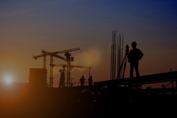 Construction workers silhouetted against the evening sky on an active building site. Cranes and steel frameworks stand in the background as workers continue tasks under fading light, suggesting extended or night-time working hours.
