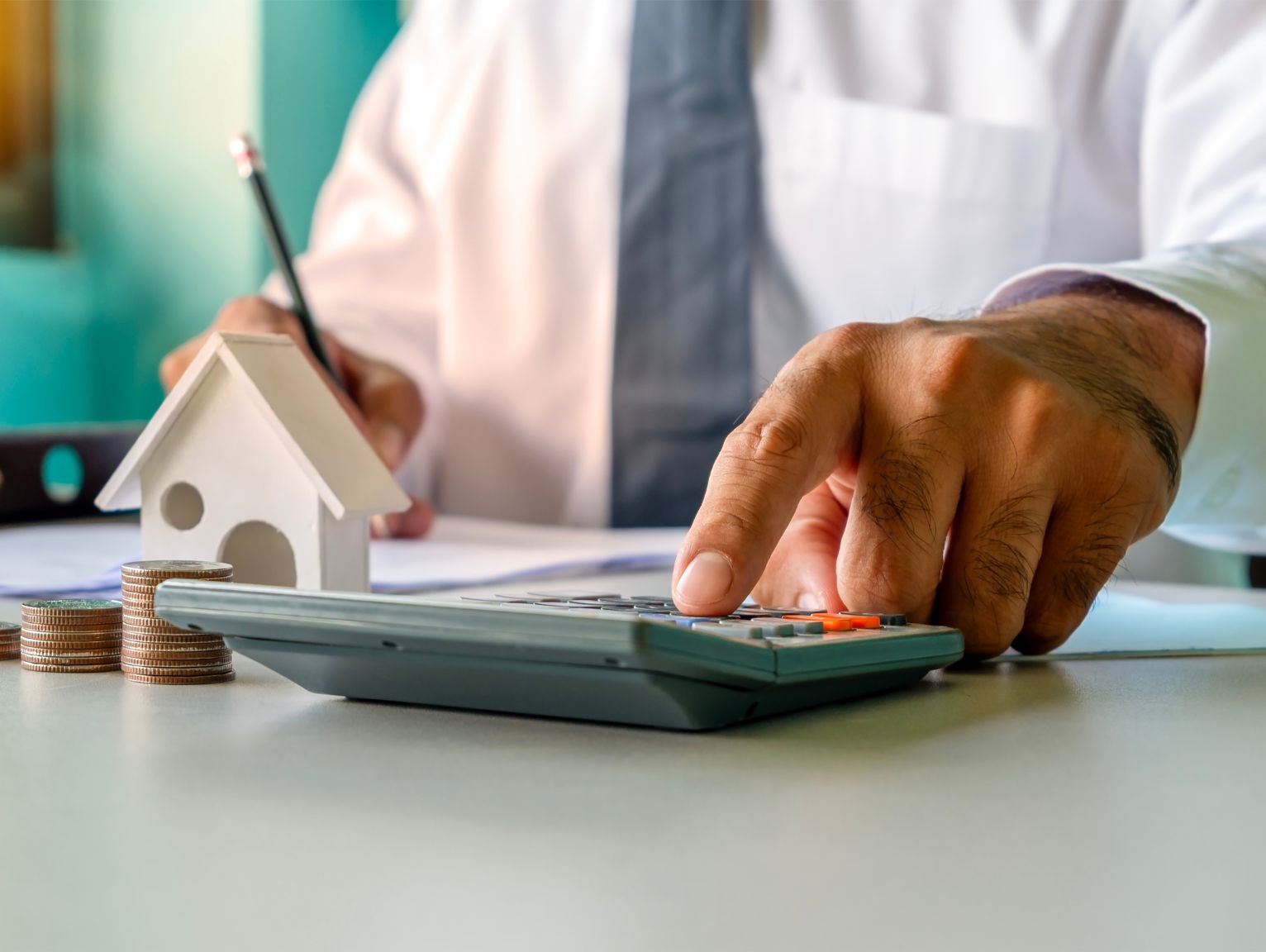 Who Pays the Building Safety Levy: A close-up of a man in a shirt and tie using a calculator and writing notes beside a small wooden house model and a stack of coins on a desk.