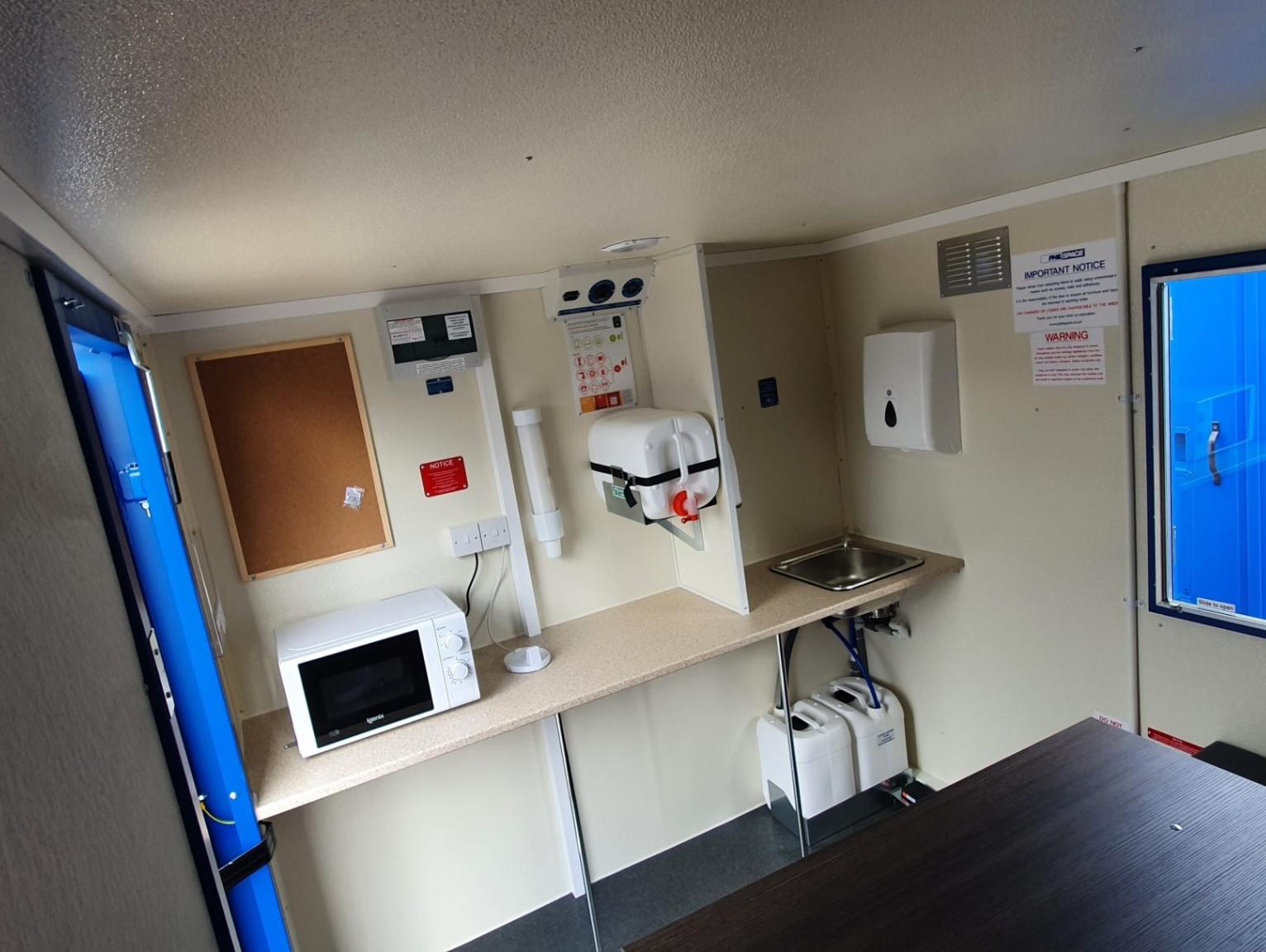 Managing welfare compliance in fit-out environments: The interior of a portable welfare unit showing a worktop with a microwave, wall-mounted hand sanitiser dispenser, stainless steel sink, paper towel dispenser, cork noticeboard, and safety signage on cream-coloured walls.