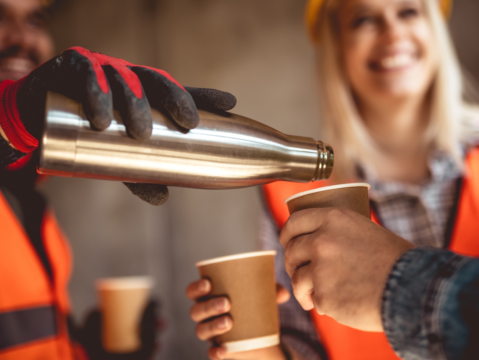 Construction workers wearing orange safety vests and gloves take a break together. One worker pours hot drinks from a metal flask into disposable cups while colleagues smile, showing a relaxed moment of teamwork during a shift.