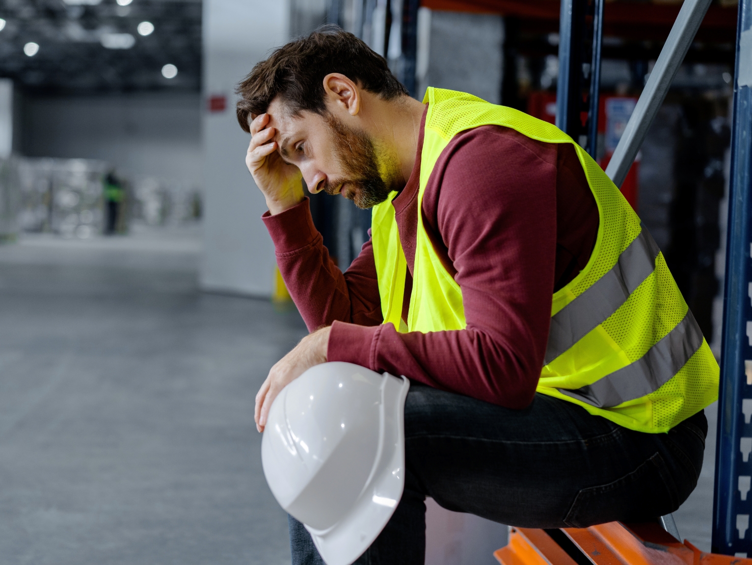 A male worker wearing a high-visibility vest sits in a warehouse, resting with his head in his hand. He looks fatigued and holds a white hard hat, conveying exhaustion or stress after a long shift.