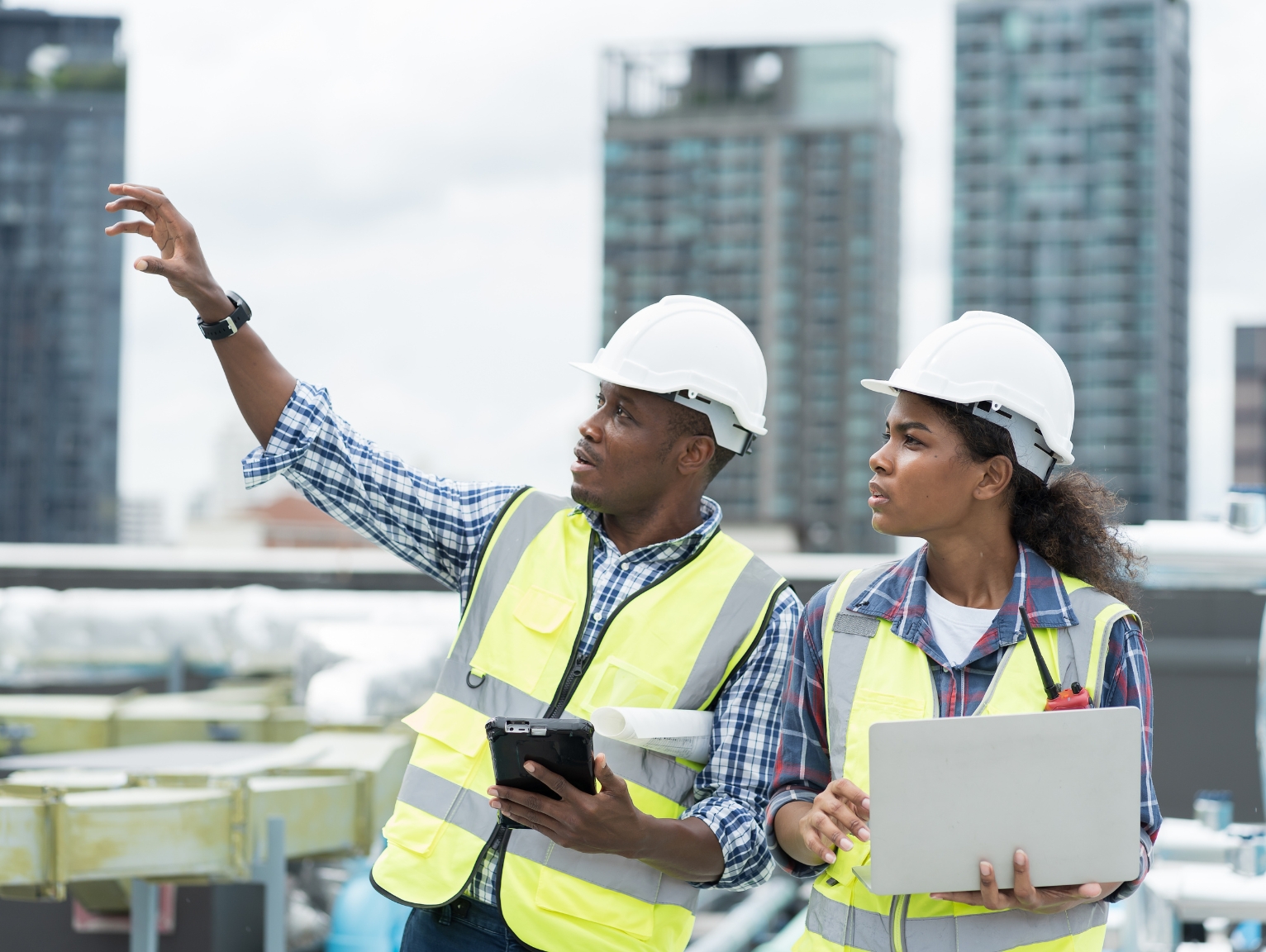 Two construction professionals in safety gear reviewing plans on a rooftop site with city buildings in the background.