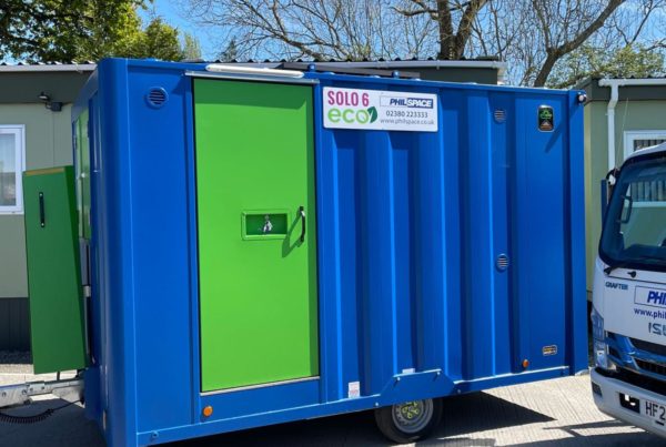 Welfare facilities for refurbishment projects: The exterior of a Philspace Solo 6 Eco portable welfare unit with a blue steel body and bright green door, photographed outdoors on a sunny day with a delivery vehicle partially visible to the right.