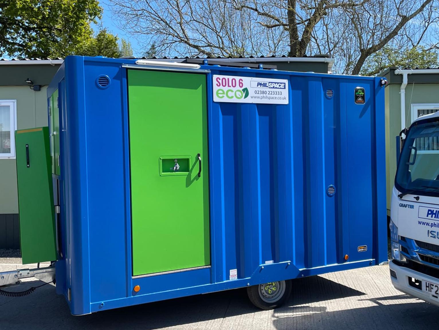 Welfare facilities for refurbishment projects: The exterior of a Philspace Solo 6 Eco portable welfare unit with a blue steel body and bright green door, photographed outdoors on a sunny day with a delivery vehicle partially visible to the right.
