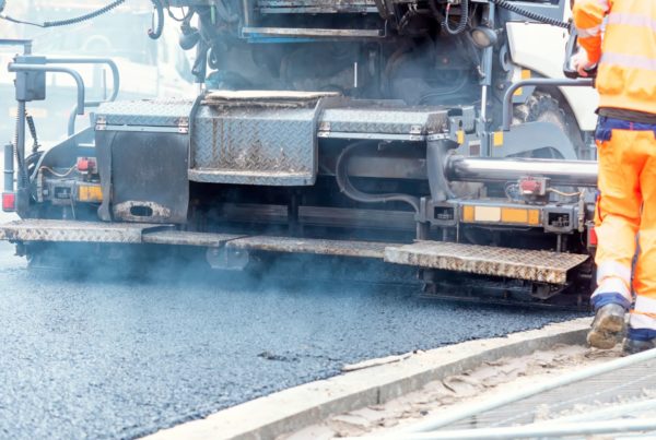 Welfare solutions for highways and utilities teams: A highways worker in a full orange high-visibility suit monitors a large asphalt paving machine laying fresh tarmac, with steam rising from the surface.