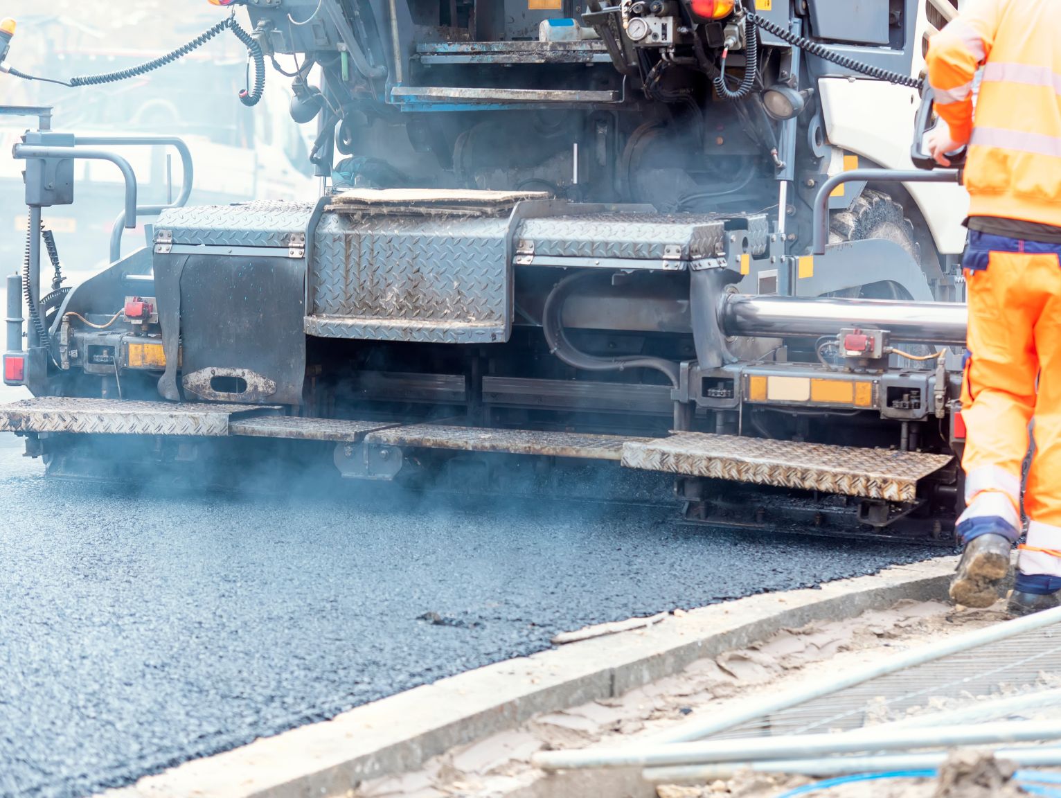 Welfare solutions for highways and utilities teams: A highways worker in a full orange high-visibility suit monitors a large asphalt paving machine laying fresh tarmac, with steam rising from the surface.