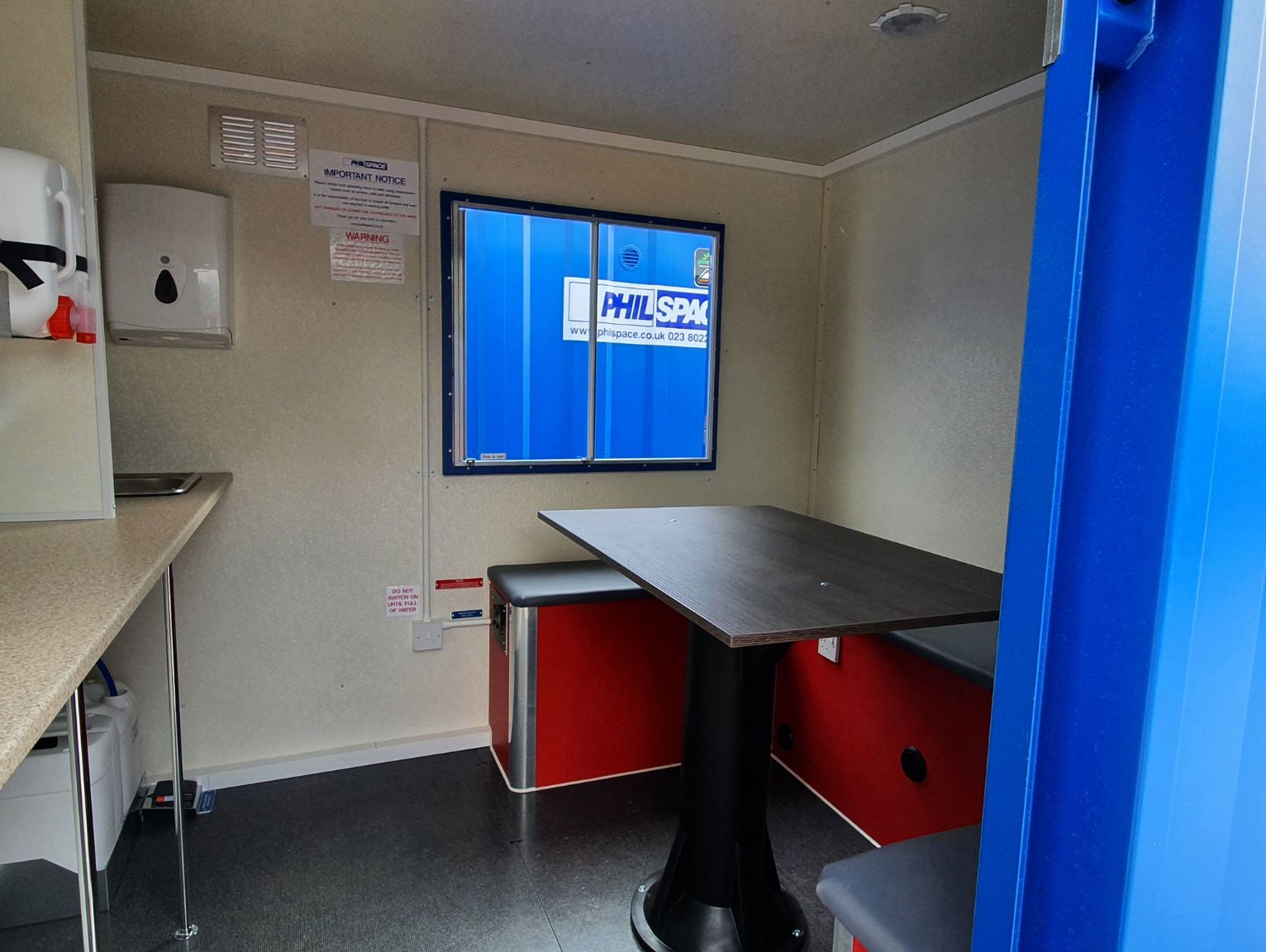 winter site setup checklist: Inside a site welfare cabin with a worktop and sink, hand towel dispenser, and a dark table with red-based bench seating beneath a window.